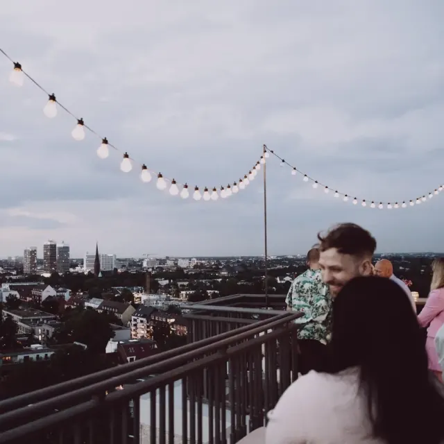 Gäste auf einer Dachterrasse mit Lichterkette über Hamburg